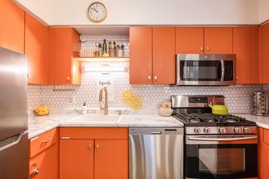 Bright modern kitchen with orange cabinets, white hexagon tile backsplash, marble countertops, brass faucet and accents, stainless steel fridge, dishwasher, microwave and gas range with a green kettle.