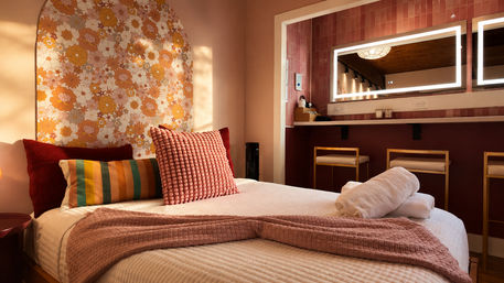 Sunlit boho-chic bedroom with retro orange-and-yellow floral arch headboard, textured pink bedspread and pillows, rolled white towels and a lighted vanity mirror with gold-framed stools.