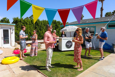 Sunny Southern California backyard summer party with adults chatting on the lawn under colorful pennant flags, palm trees, inflatable pool rings and a small outdoor drink cart