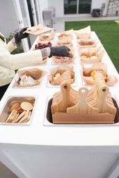 Outdoor patio charcuterie station with paper-lined trays of cheese cubes, crackers, grapes and snacks, stacked mini wooden cutting boards, and gloved hands arranging servings