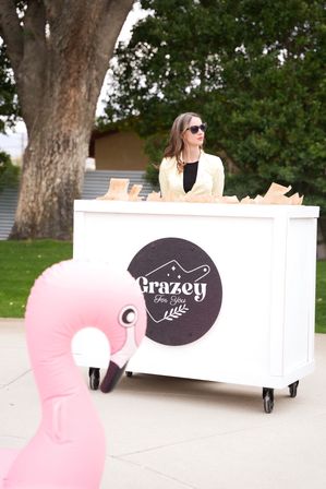 Woman wearing sunglasses behind a white mobile vendor cart at a park, paper bags on the counter and a bright inflatable pink flamingo in the foreground