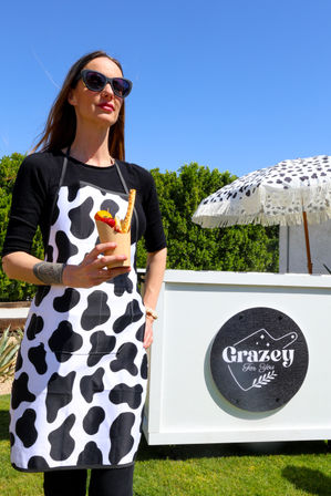 Woman in black-and-white cow-print apron and sunglasses holding a fruit-topped dessert cone at a sunny outdoor dessert cart with a fringed umbrella