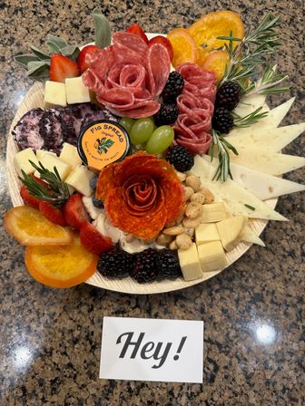 Charcuterie board on a granite countertop with salami and pepperoni carved into roses, assorted sliced and cubed cheeses, blackberries, strawberries, green grapes, candied orange slices, nuts, a small jar of fig spread, rosemary sprigs, and a small card reading 'Hey!'