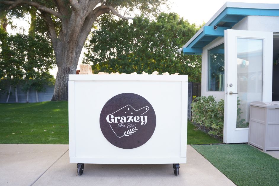 White portable serving cart with round black logo on front, sitting on a sunlit backyard patio beside an open glass door and green lawn under a large shade tree.