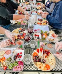 Al fresco charcuterie and wine tasting on a metal patio table with multiple boards of cheese, salami, grapes, berries, crackers, nuts, wine glasses and gloved hands arranging plates.