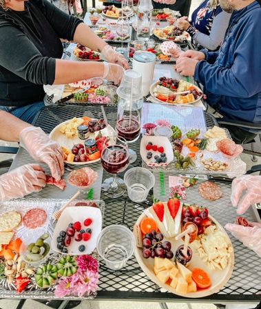 Al fresco charcuterie and wine tasting on a metal patio table with multiple boards of cheese, salami, grapes, berries, crackers, nuts, wine glasses and gloved hands arranging plates.