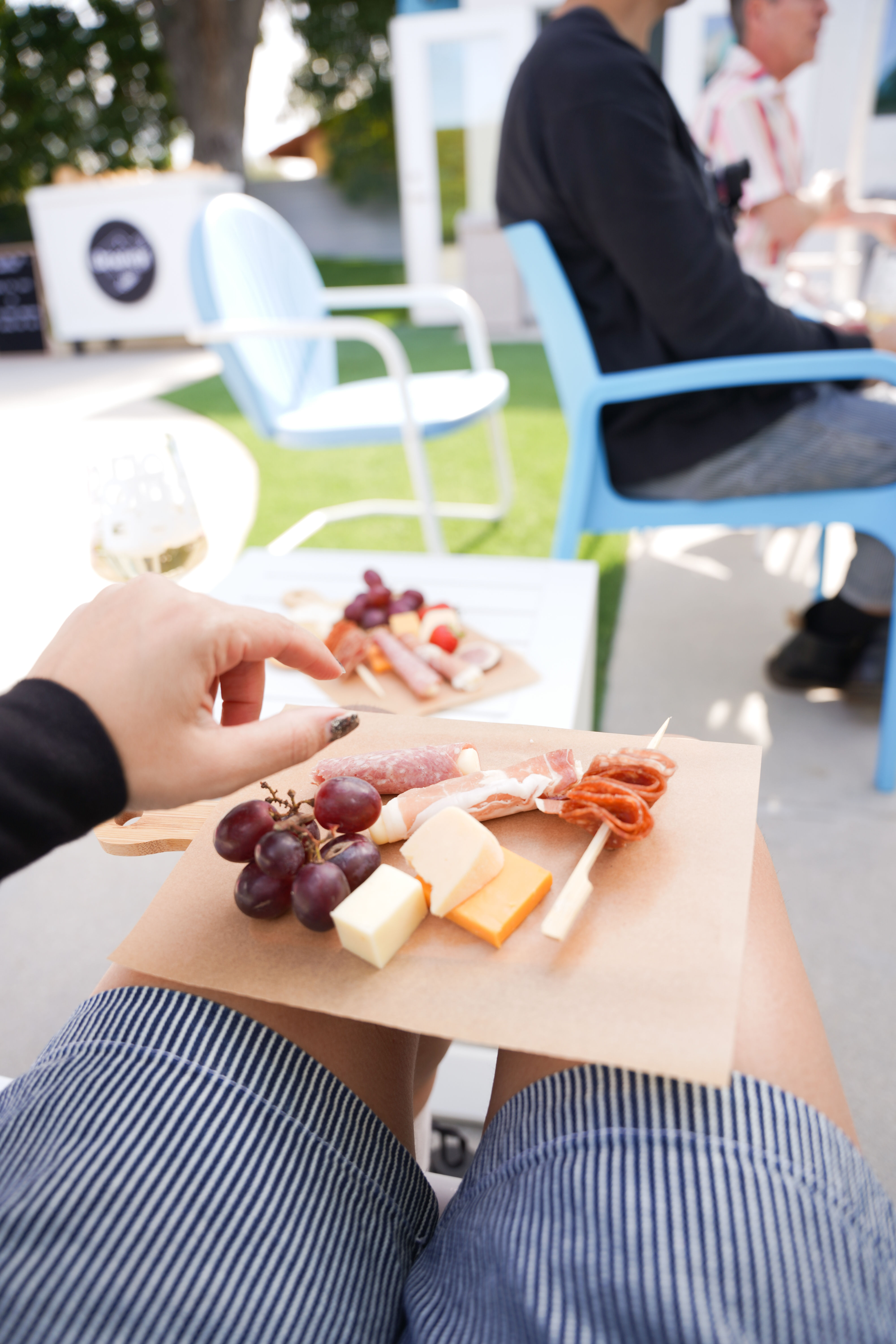 Mini charcuterie board with grapes, cheese cubes and cured meats on a lap at a sunny outdoor patio with blue chairs and a glass of white wine