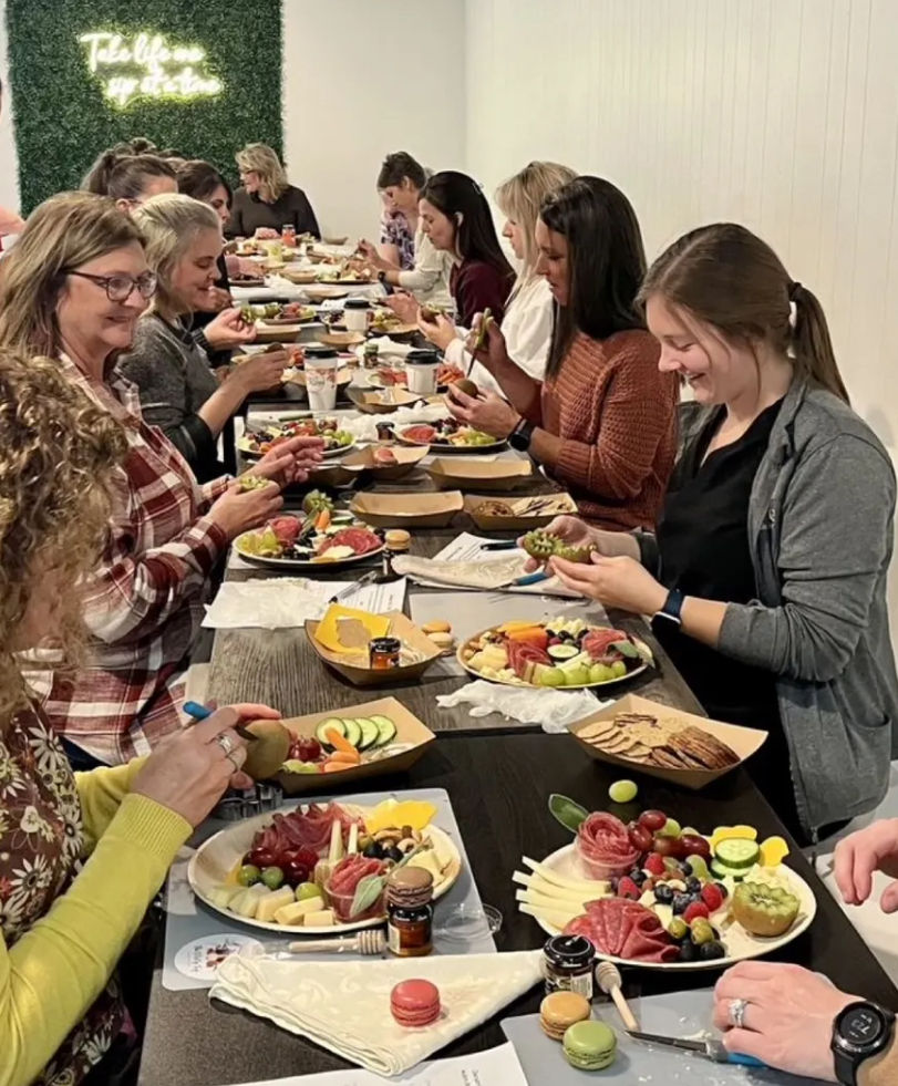 Group charcuterie board workshop with people seated at a long table assembling colorful fruit, cheese, crackers and macarons in an indoor hands-on class.