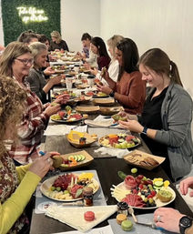 Group charcuterie board workshop with people seated at a long table assembling colorful fruit, cheese, crackers and macarons in an indoor hands-on class.