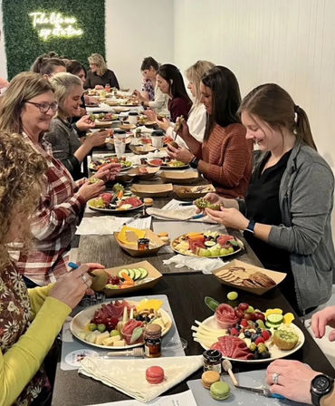 Group charcuterie board workshop with people seated at a long table assembling colorful fruit, cheese, crackers and macarons in an indoor hands-on class.