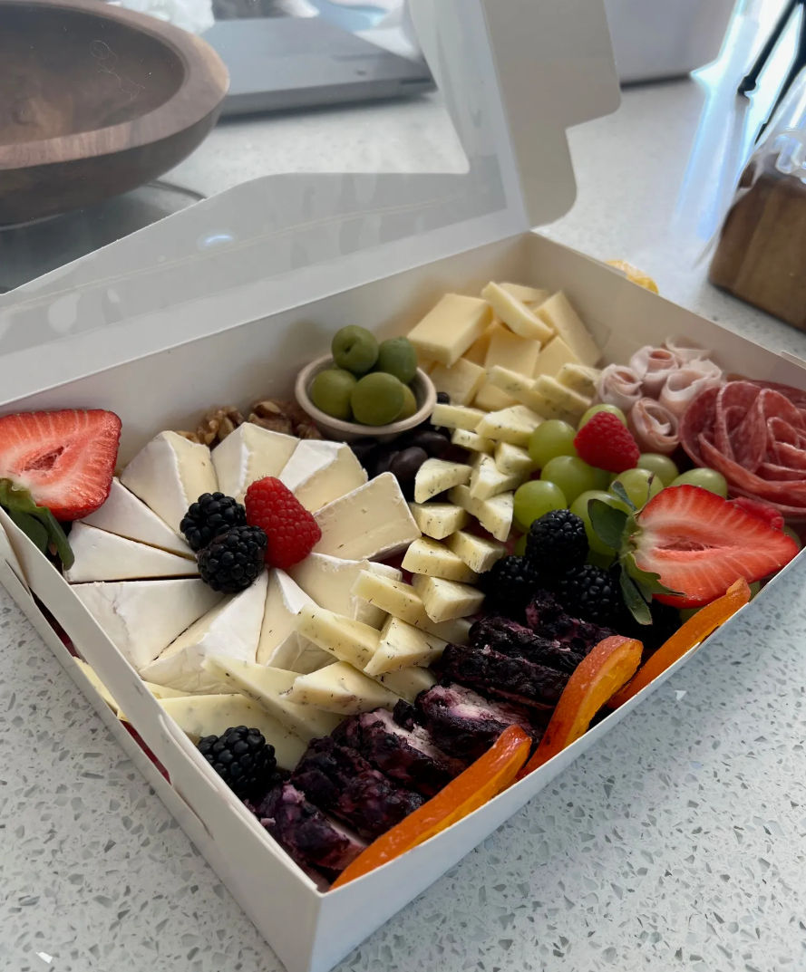 Gourmet charcuterie box on a kitchen counter with a sliced wheel of brie, cubed semi-hard cheeses, blueberry-marble cheese, rolled cured meats, green grapes, strawberries, blackberries, raspberries, green olives, walnuts and dried apricot slices — perfect picnic or party snack.