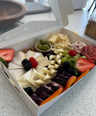 Gourmet charcuterie box on a kitchen counter with a sliced wheel of brie, cubed semi-hard cheeses, blueberry-marble cheese, rolled cured meats, green grapes, strawberries, blackberries, raspberries, green olives, walnuts and dried apricot slices — perfect picnic or party snack.