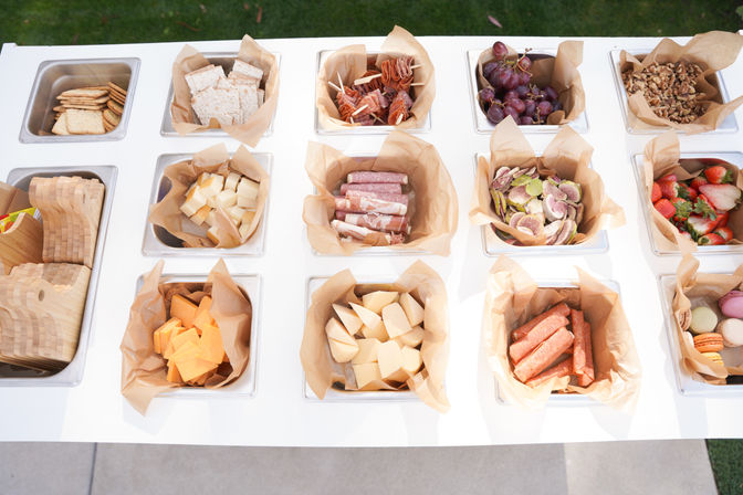 Top-down view of an outdoor charcuterie buffet on a white table with parchment-lined trays holding crackers, assorted cheeses, cured meats, grapes, strawberries, nuts and colorful macarons.
