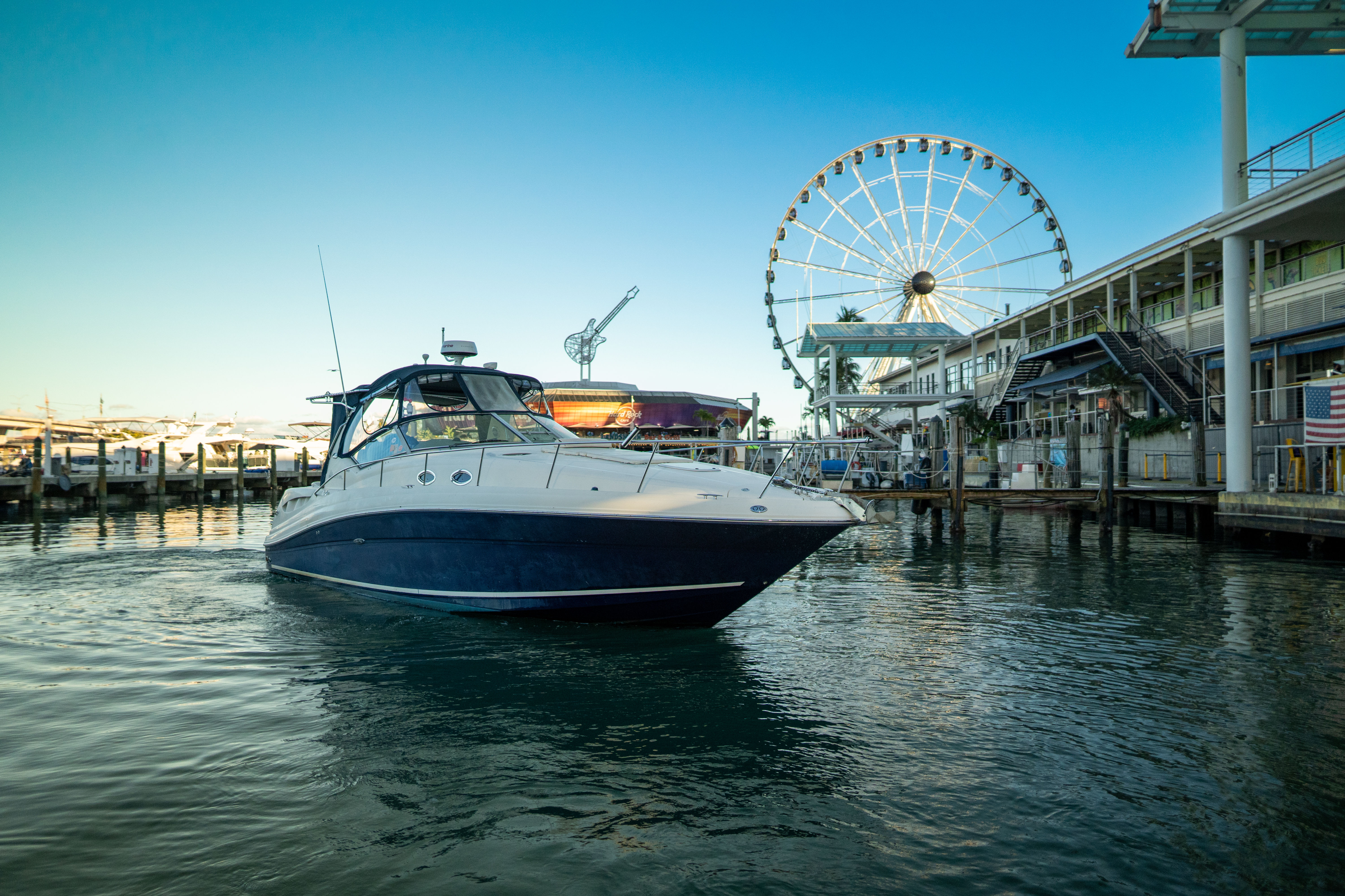 Blue-and-white motor yacht in a calm waterfront marina with a large Ferris wheel, piers and seaside buildings under a clear sky.