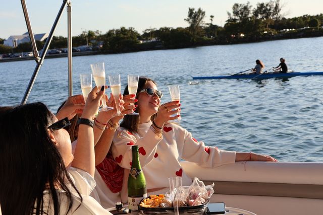 Sunlit group on a boat toasting with champagne flutes over a coastal bay, snacks on the table and a rowing team passing in the background.