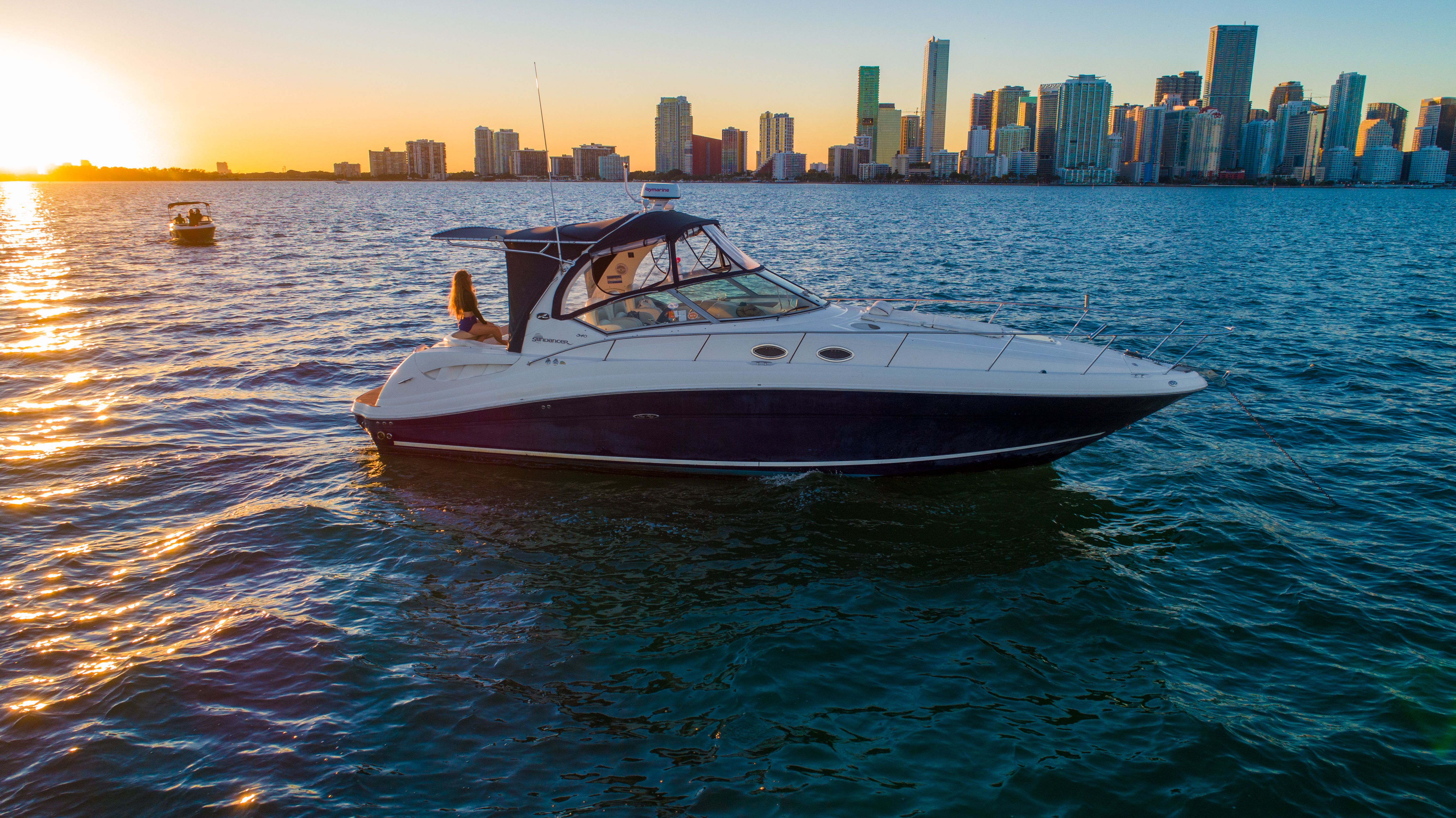Luxury motor yacht on shimmering water with a person on the stern, set against an urban high-rise skyline at sunset.