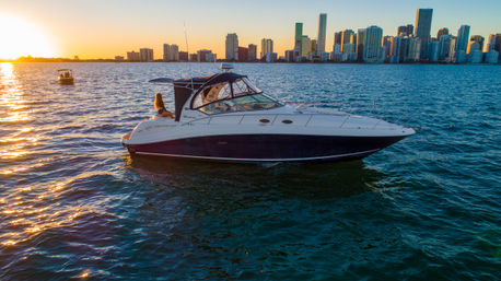 Luxury motor yacht on shimmering water with a person on the stern, set against an urban high-rise skyline at sunset.