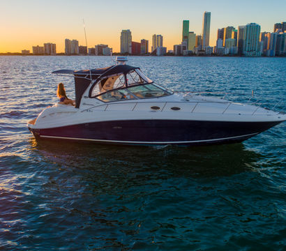 Luxury motor yacht on shimmering water with a person on the stern, set against an urban high-rise skyline at sunset.