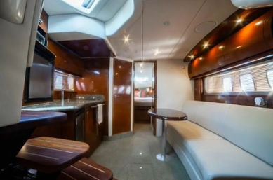 Sleek luxury yacht cabin interior: glossy wood-paneled galley with granite countertop and fridge, curved cream leather sofa with round table, skylight and doorway to sleeping cabin.