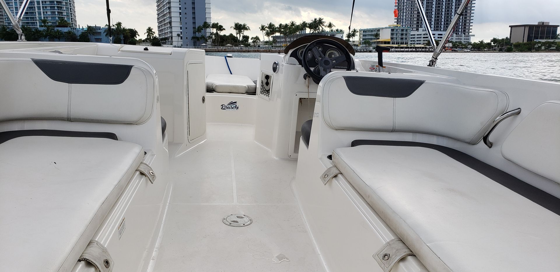 White motorboat interior with cushioned bench seats and helm, facing a palm‑lined waterfront and high‑rise condos under a cloudy sky