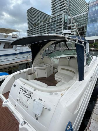 White luxury cabin cruiser docked at a marina with cushioned cockpit seating, navy canvas bimini, teak deck, and waterfront high‑rise buildings in the background.