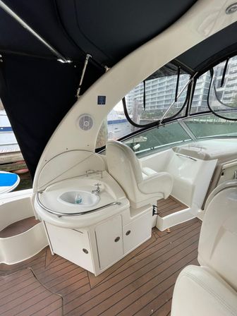Yacht cockpit at a marina with a white wet bar sink and storage cabinet, cushioned helm seat, synthetic teak-style deck and black canvas enclosure, waterfront high-rise condos visible through the windows.