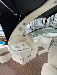 Boat cockpit with cream captain’s chair, built-in wet sink and storage cabinets, teak-style deck, black canvas bimini and marina buildings visible through vinyl windows — cozy yacht helm area ready for cruising.