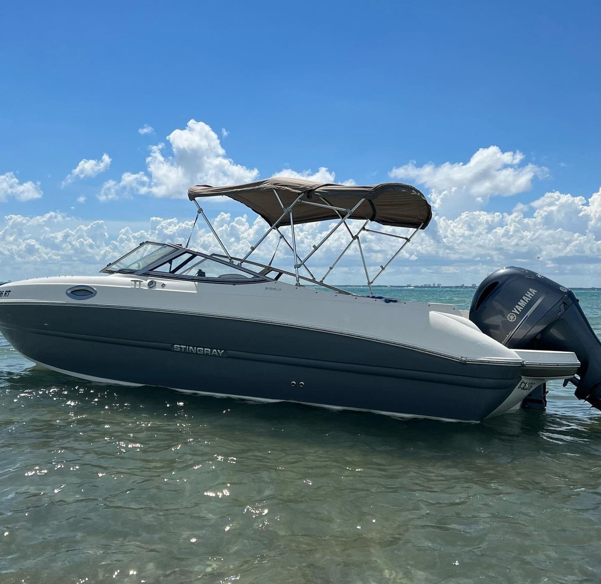 Sleek gray-and-white motorboat with tan bimini top floating in shallow clear turquoise coastal water under a bright blue sky dotted with puffy white clouds