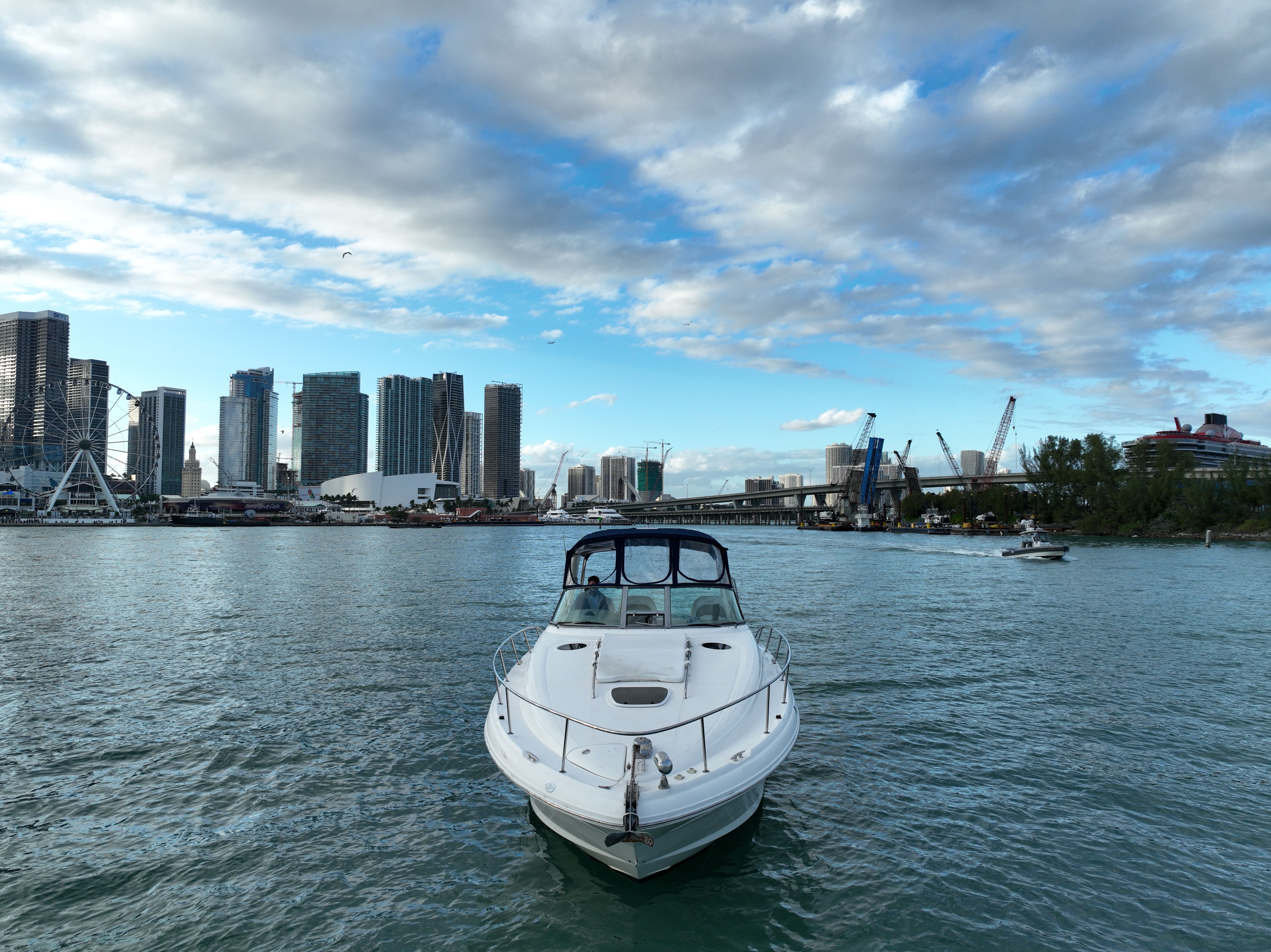 White motorboat approaching on the Miami waterfront with downtown skyline, Ferris wheel, bridge and a cruise ship under a partly cloudy blue sky