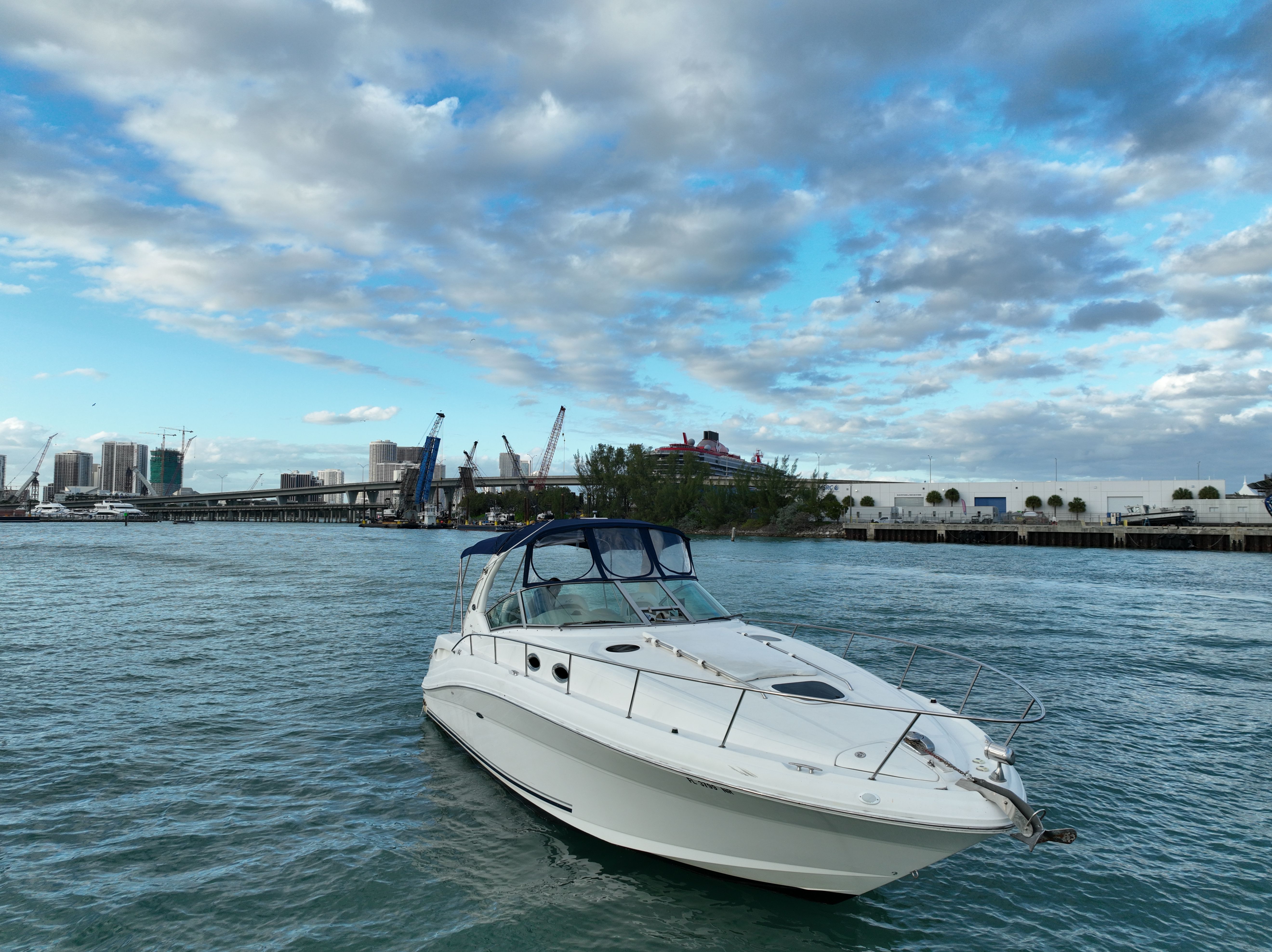 Sleek white motor yacht floating in a busy urban harbor, with waterfront skyline, cranes and bridge under a dramatic cloud-streaked blue sky
