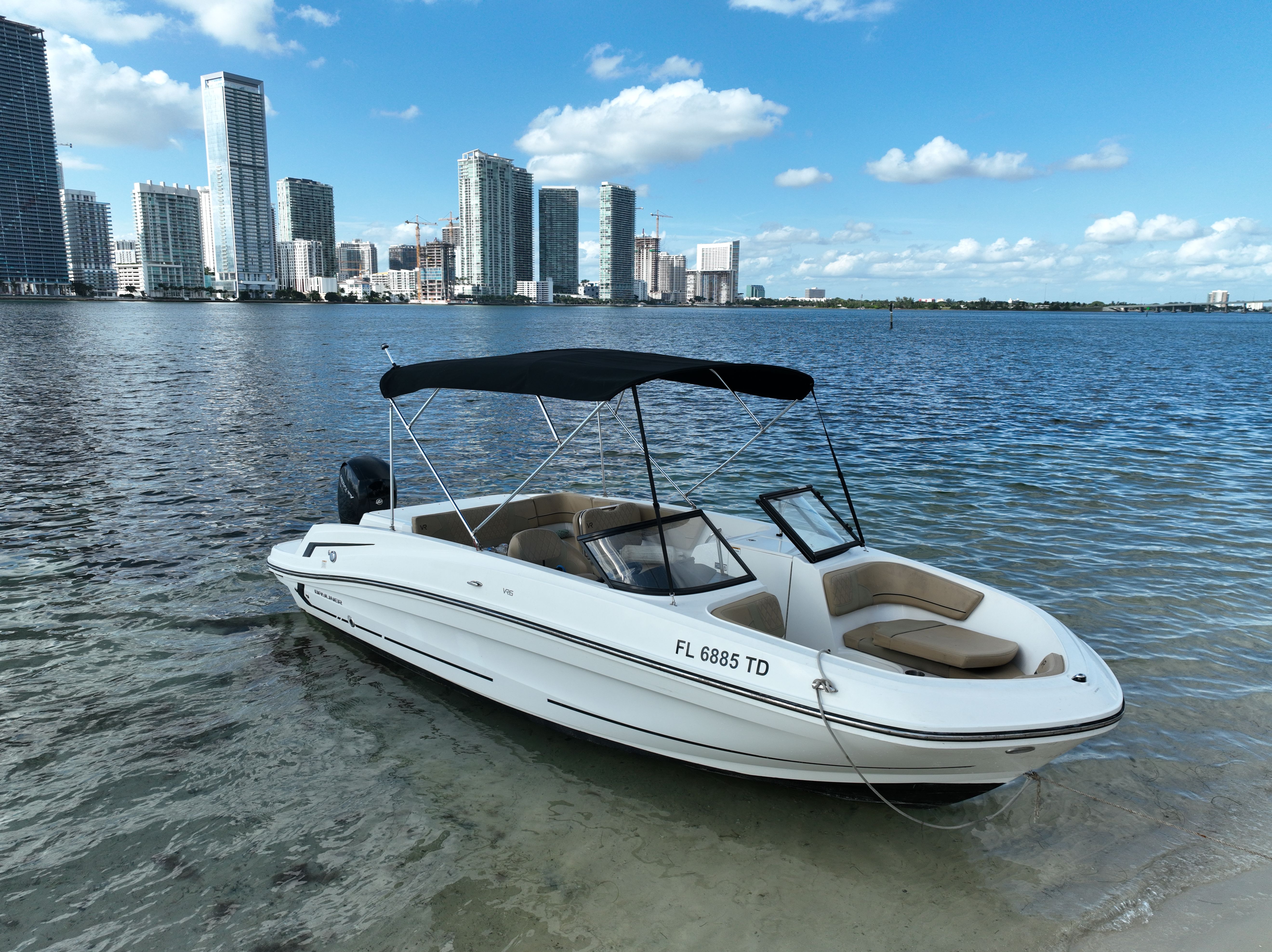 White bowrider motorboat with tan cushions and black bimini anchored in clear shallow Florida bay water, with a modern high‑rise skyline and blue sky with clouds in the background.