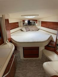 Cozy yacht interior V-berth cabin with rounded queen bed, patterned bedding and pillows, wood storage cabinets, small galley counter and cushioned seating under a ceiling hatch.