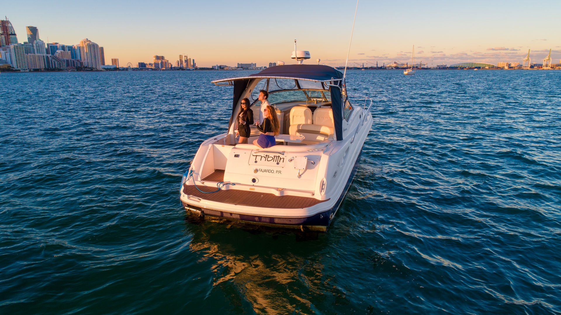 White motor yacht with three people on the aft deck cruising on blue water near an urban skyline at golden-hour sunset
