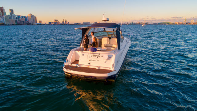 White motor yacht with three people on the aft deck cruising on blue water near an urban skyline at golden-hour sunset