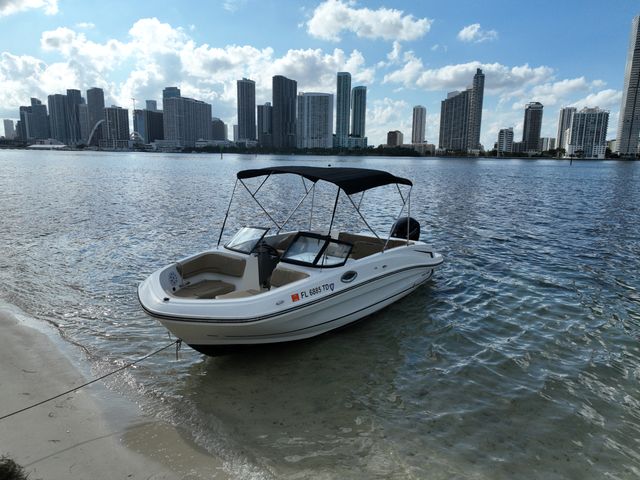 White motorboat with black canopy anchored at a sandy Florida waterfront, high-rise city skyline across the bay under a bright blue sky.