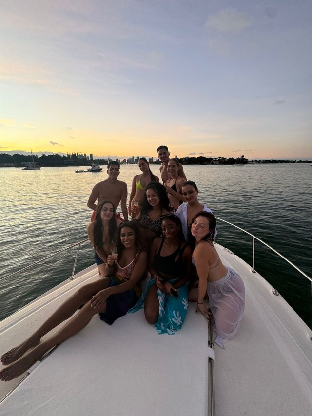 Group of friends on a yacht bow at sunset during a calm bay cruise, smiling and holding drinks with a coastal city skyline on the horizon.