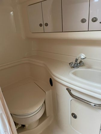 Compact cream fiberglass boat head with molded sink and chrome faucet, closed toilet, overhead storage cabinets and stainless grab rail