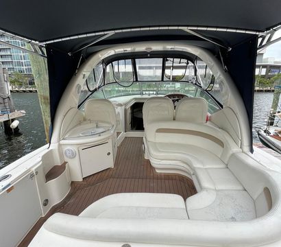 Cozy motor yacht cockpit with curved cream vinyl lounge seating and teak-style deck beneath a navy bimini top, small wet bar and helm visible, docked at a marina with waterfront buildings
