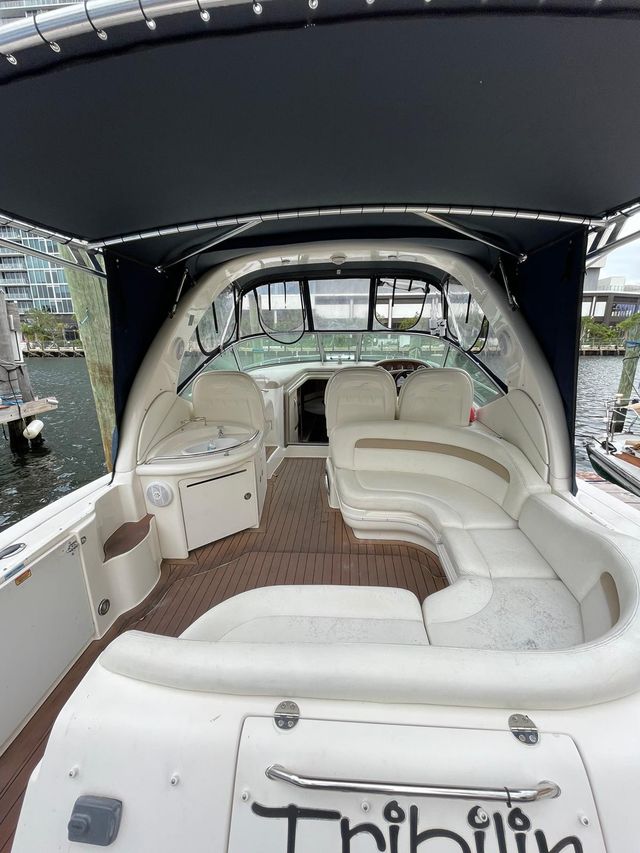 Cozy motor yacht cockpit with curved cream vinyl lounge seating and teak-style deck beneath a navy bimini top, small wet bar and helm visible, docked at a marina with waterfront buildings