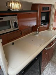 Compact yacht galley: creamy solid-surface countertop with covered sink and curved faucet, built-in microwave/coffee unit, glossy cherry wood cabinets and small curtained porthole.
