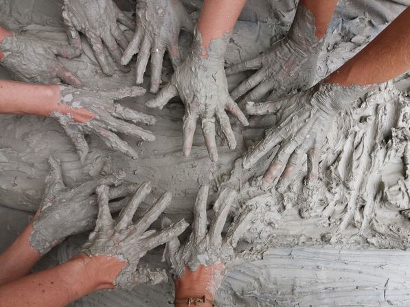 Several hands covered in wet gray clay pressing and shaping a large slab during a group pottery workshop