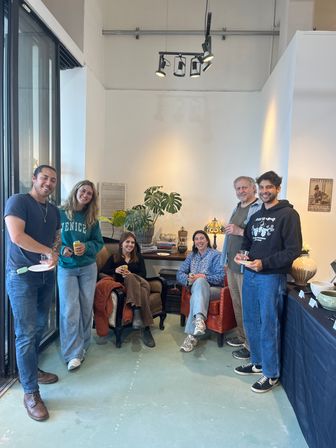 Six people smiling in a light-filled gallery-style studio, standing and sitting around leather armchairs and a side table with a large monstera plant and pottery, holding drinks and plates under black track lighting.