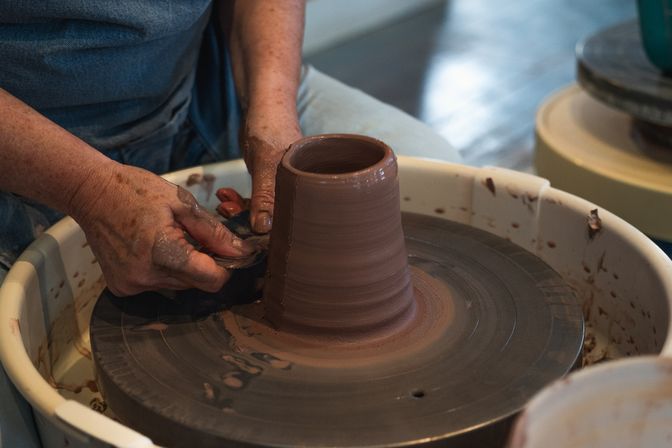 Hands shaping a tall brown clay cup on a spinning potter's wheel, trimming wet clay with a metal tool in a pottery studio.
