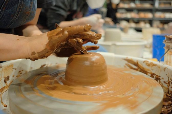 Close-up of clay-covered hands shaping a spinning brown pot on a pottery wheel in a ceramics studio.