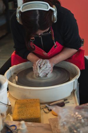 Close-up of a potter shaping wet clay on a spinning pottery wheel in a ceramic studio, hands covered in clay, wearing a red apron and headphones with tools and sponge nearby.
