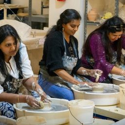 Three women in a group pottery class shaping clay on pottery wheels in a ceramics studio, hands and aprons covered in clay with buckets nearby.