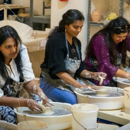 Three women in a group pottery class shaping clay on pottery wheels in a ceramics studio, hands and aprons covered in clay with buckets nearby.