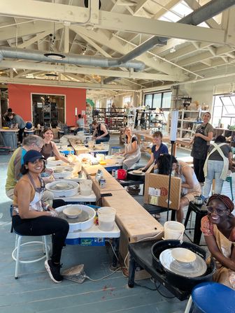 Community pottery class with smiling students at pottery wheels in a bright loft-style ceramics studio, shaping clay bowls and plates amid shelving of supplies and natural light