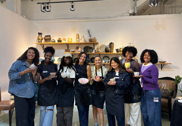 Nine women in aprons smiling and proudly holding handmade ceramic bowls and cups in a pottery studio workshop with shelves of finished pottery behind them.
