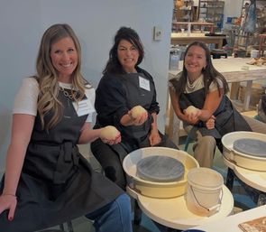 Three people in black aprons smiling and holding balls of clay while seated at pottery wheels in a bright ceramics studio, hands-on wheel-throwing class with buckets and tools visible